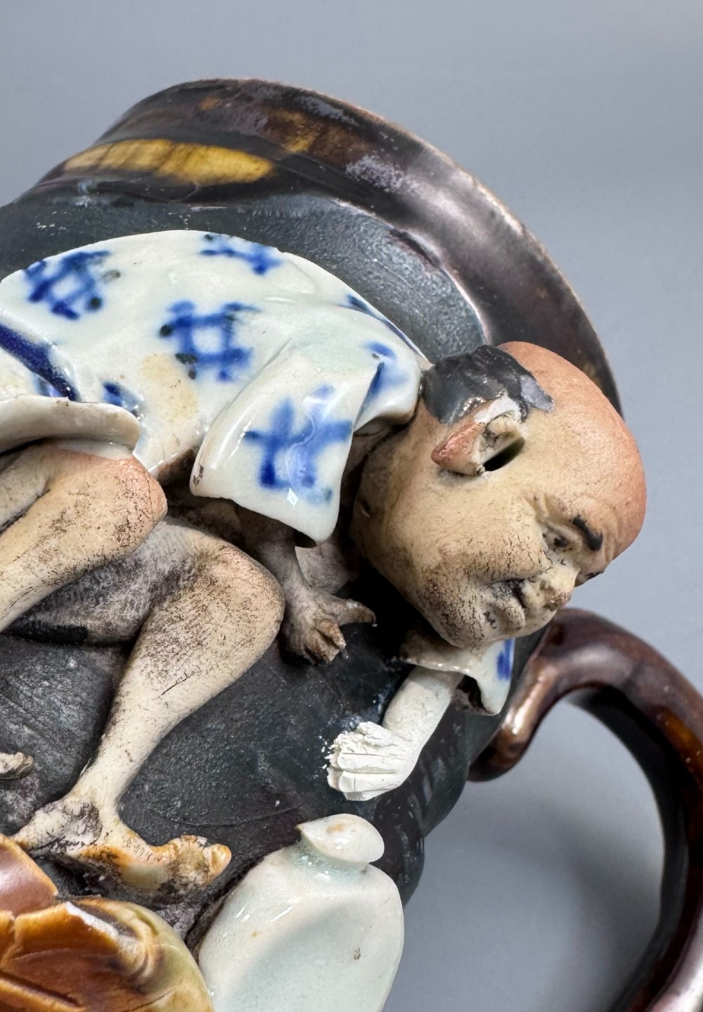 A dark ribbed ceramic mug featuring a dripping flambe glaze rim and an applied relief figure of a seated Japanese sennin. View XII