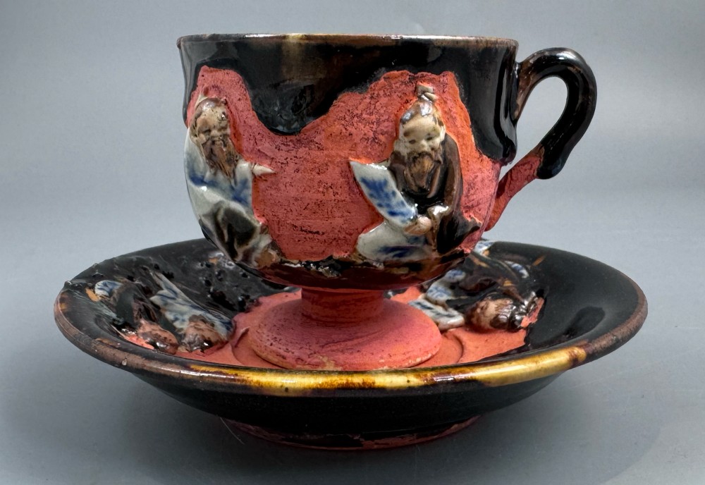 A Japanese Sumida Gawa teacup and saucer featuring relief-carved sage figures on a textured red background with a heavy dark flambe glaze.