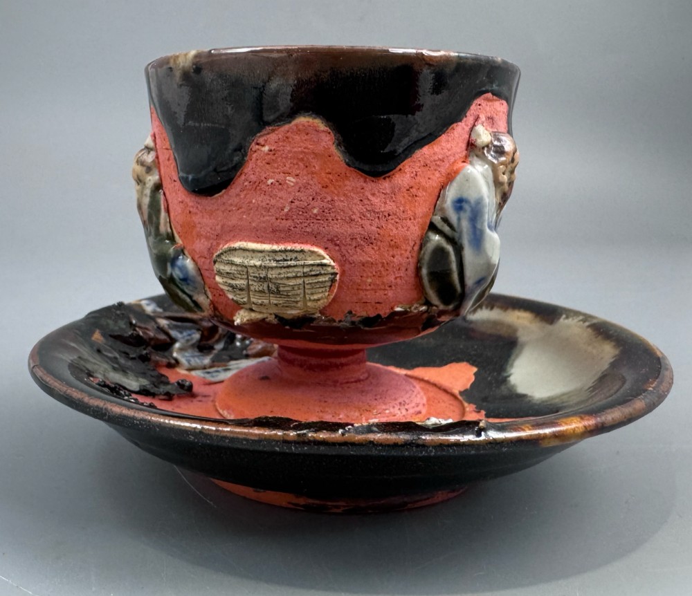 A Japanese Sumida Gawa teacup and saucer featuring relief-carved sage figures on a textured red background with a heavy dark flambe glaze. View II