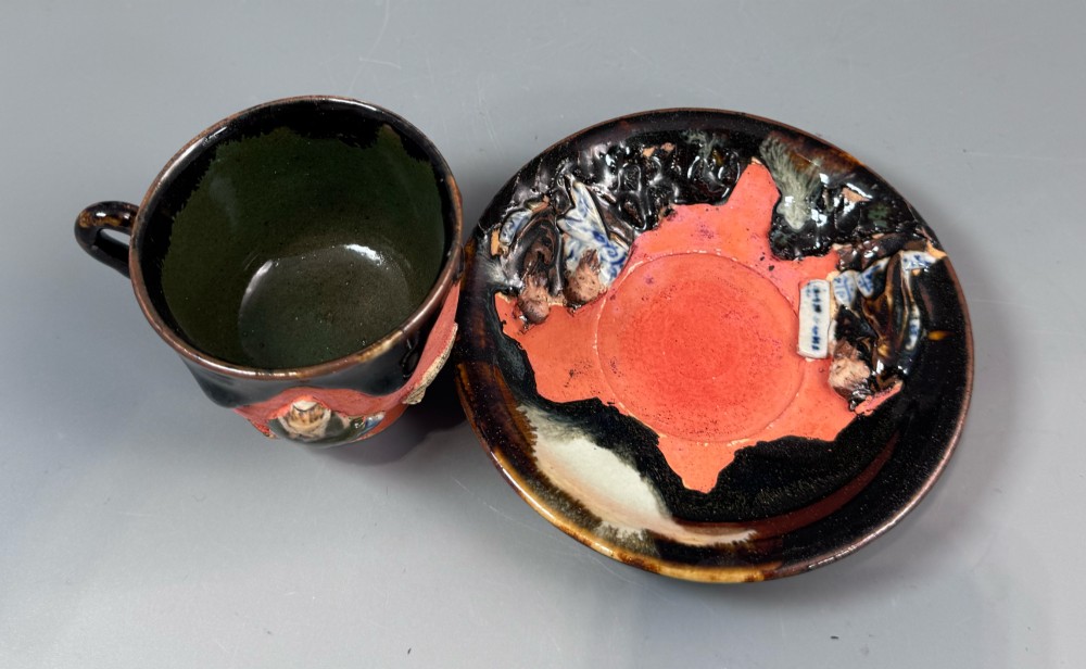 A Japanese Sumida Gawa teacup and saucer featuring relief-carved sage figures on a textured red background with a heavy dark flambe glaze. View IV