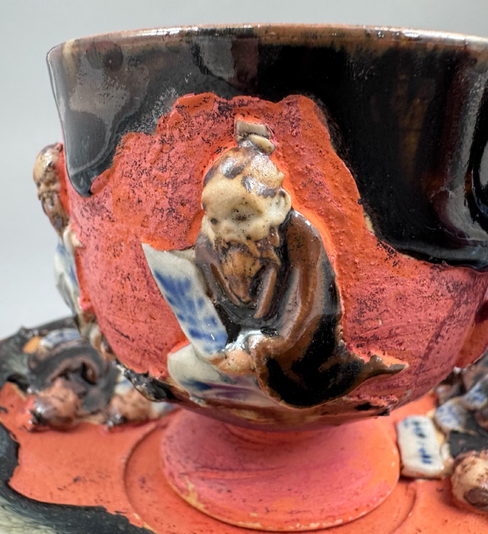 A Japanese Sumida Gawa teacup and saucer featuring relief-carved sage figures on a textured red background with a heavy dark flambe glaze. View V