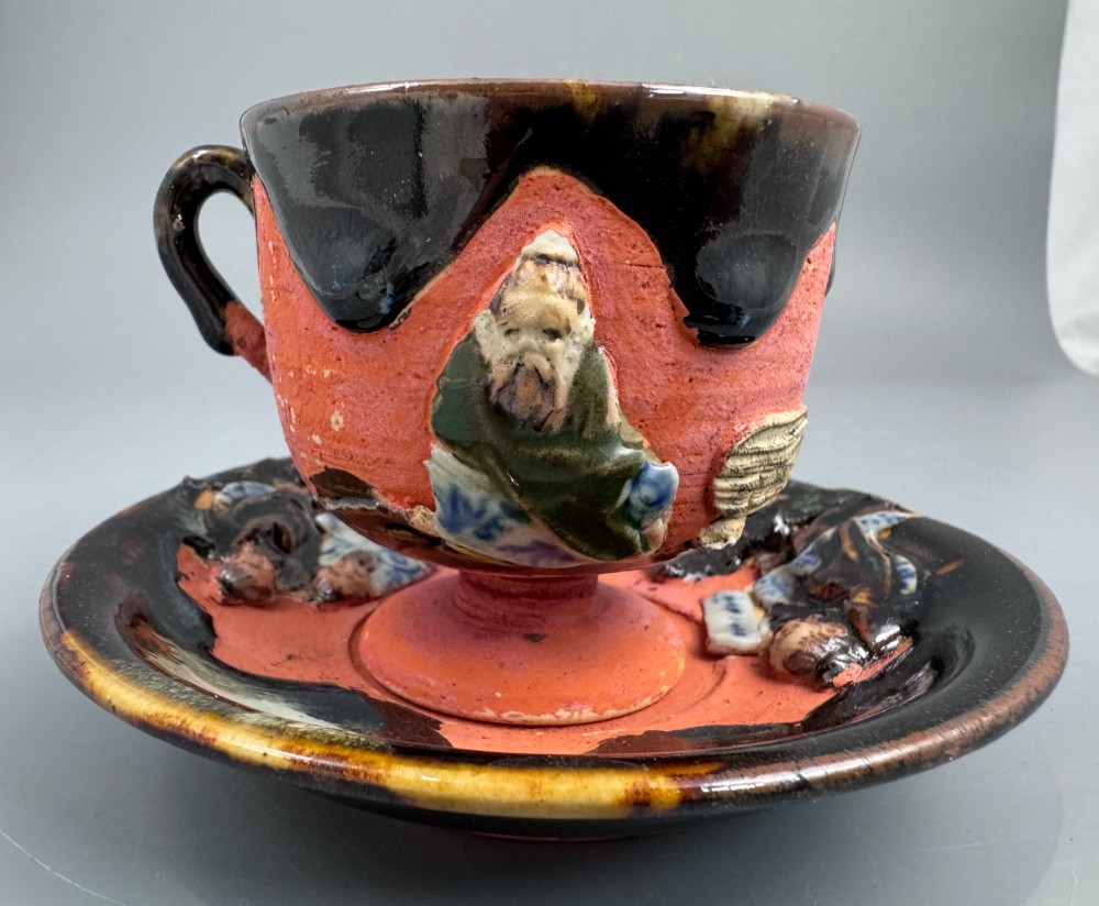 A Japanese Sumida Gawa teacup and saucer featuring relief-carved sage figures on a textured red background with a heavy dark flambe glaze. View VI