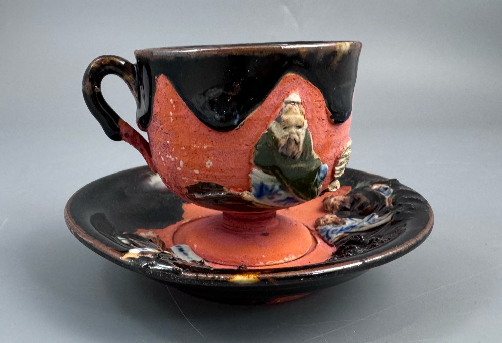 A Japanese Sumida Gawa teacup and saucer featuring relief-carved sage figures on a textured red background with a heavy dark flambe glaze. View XII