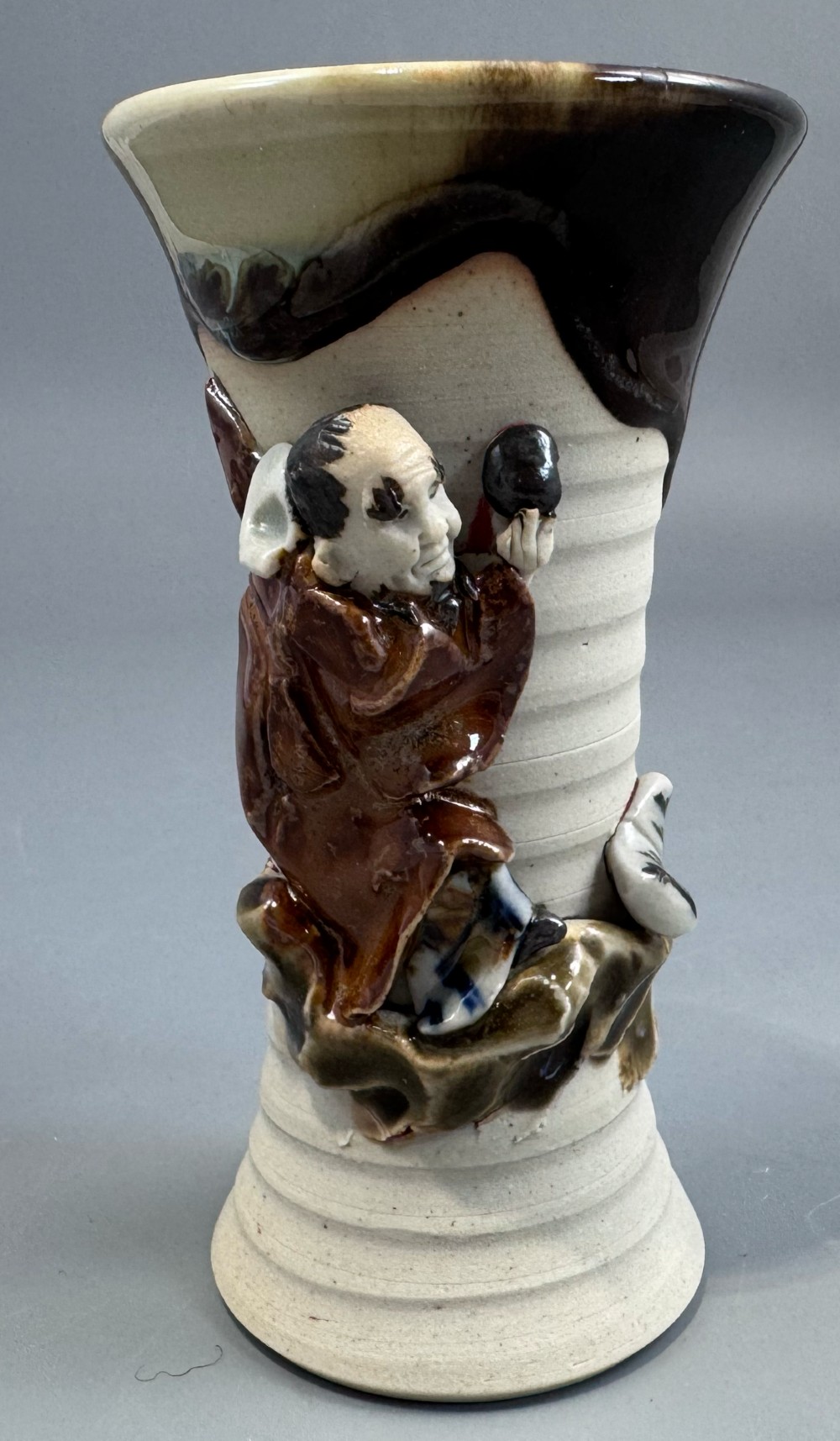 A Japanese Sumida Gawa stoneware vase featuring a ribbed cream body, dripping dark glaze along the flared rim, and an applied high-relief figure of a Rakan. View III