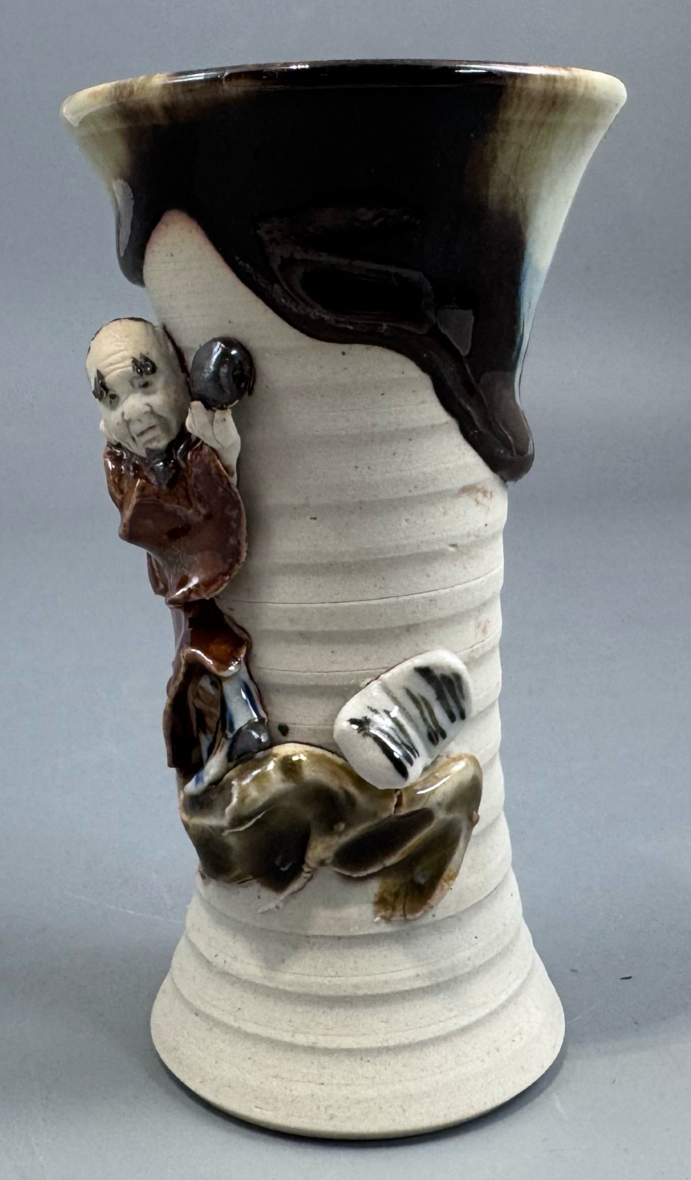 A Japanese Sumida Gawa stoneware vase featuring a ribbed cream body, dripping dark glaze along the flared rim, and an applied high-relief figure of a Rakan. View IV