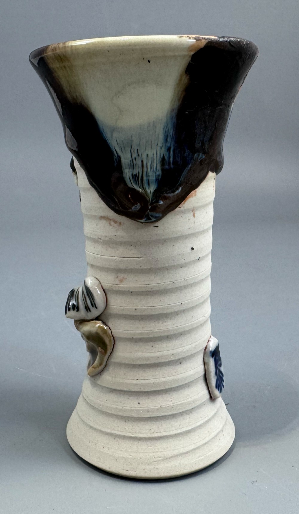 A Japanese Sumida Gawa stoneware vase featuring a ribbed cream body, dripping dark glaze along the flared rim, and an applied high-relief figure of a Rakan. View V