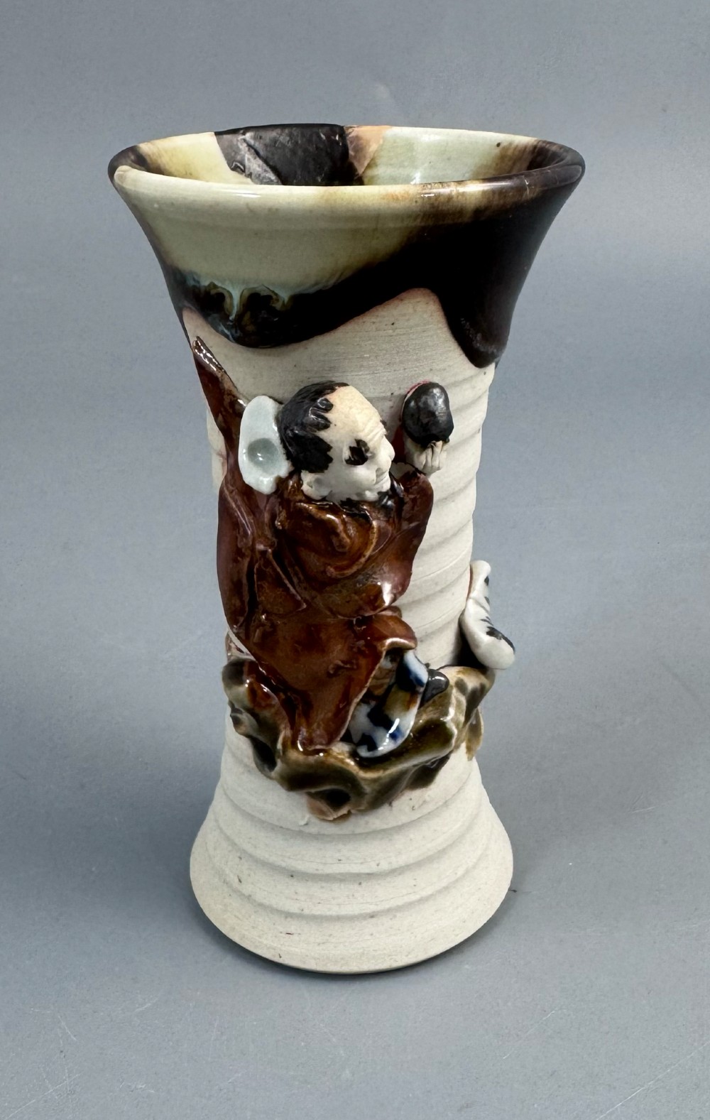 A Japanese Sumida Gawa stoneware vase featuring a ribbed cream body, dripping dark glaze along the flared rim, and an applied high-relief figure of a Rakan. View VII