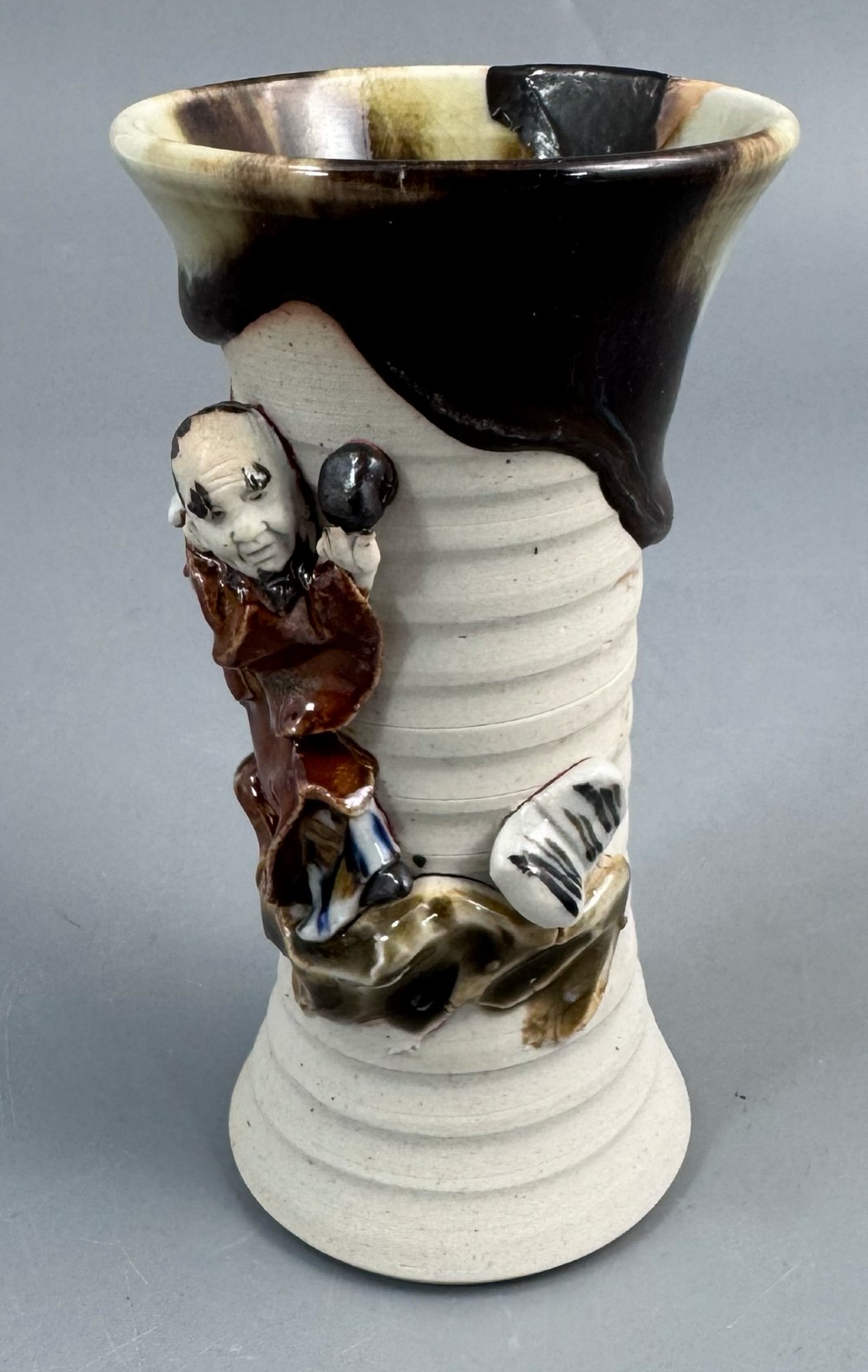 A Japanese Sumida Gawa stoneware vase featuring a ribbed cream body, dripping dark glaze along the flared rim, and an applied high-relief figure of a Rakan. View VIII