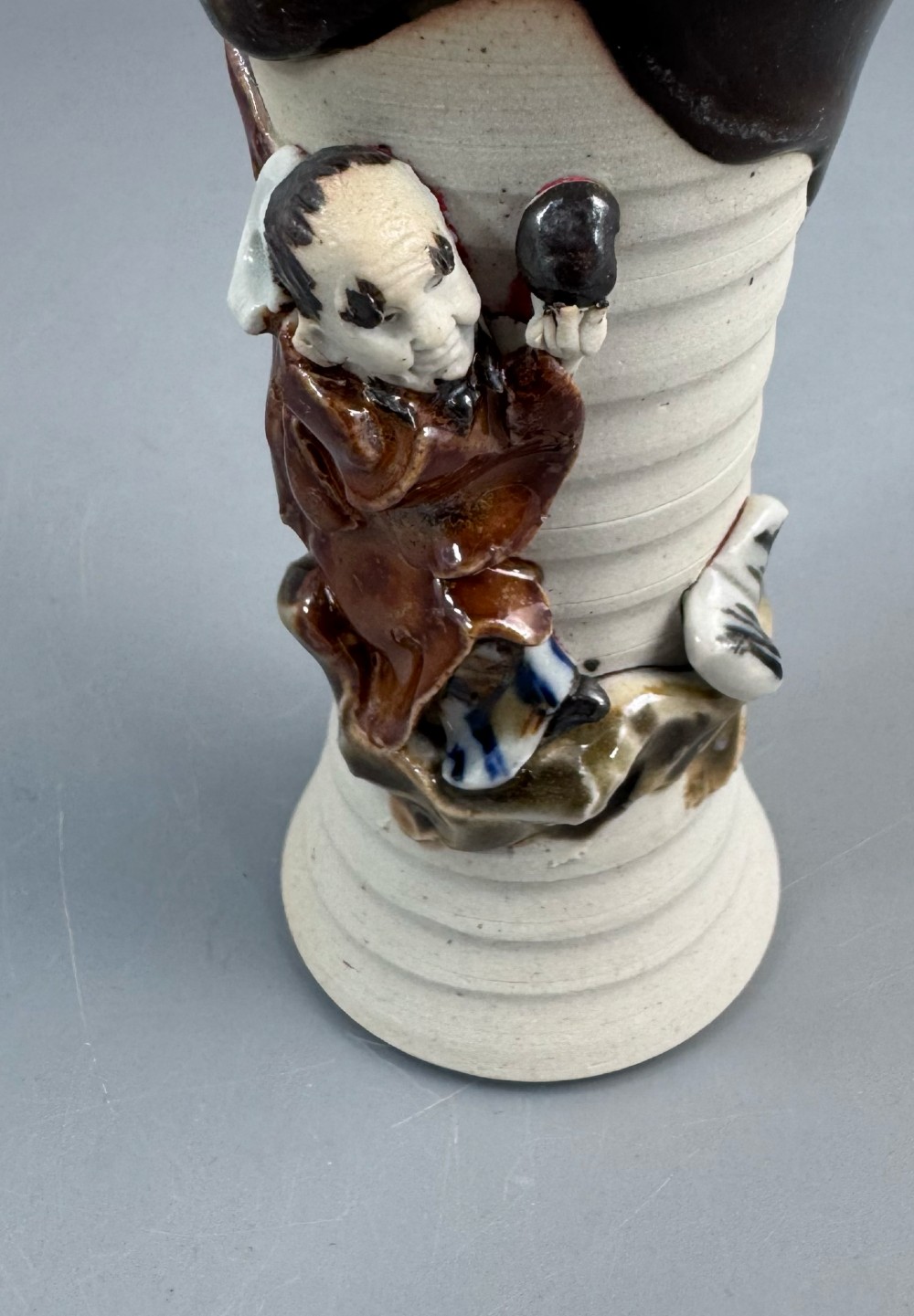 A Japanese Sumida Gawa stoneware vase featuring a ribbed cream body, dripping dark glaze along the flared rim, and an applied high-relief figure of a Rakan. View X