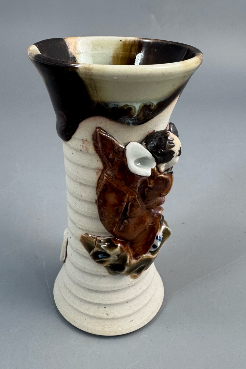 A Japanese Sumida Gawa stoneware vase featuring a ribbed cream body, dripping dark glaze along the flared rim, and an applied high-relief figure of a Rakan. View XV