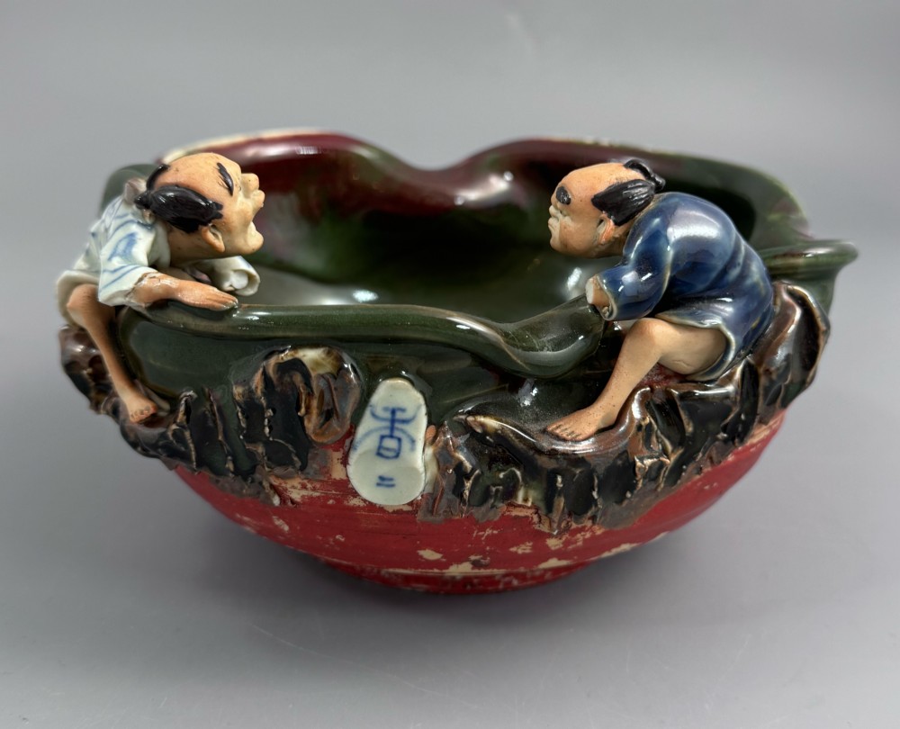 A Japanese Sumida Gawa pottery bowl featuring a dark green running glaze over a red base with two sculpted figures leaning over the rim and an applied white makers mark.