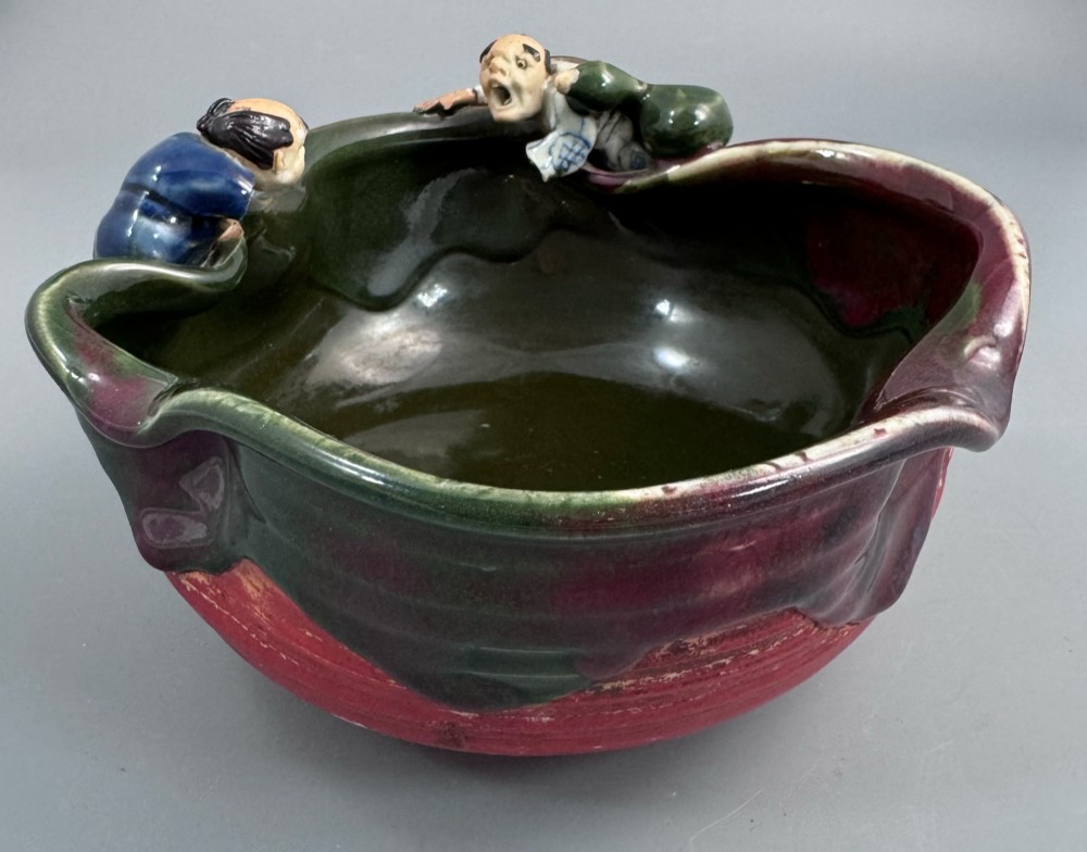 A Japanese Sumida Gawa pottery bowl featuring a dark green running glaze over a red base with two sculpted figures leaning over the rim and an applied white makers mark. View IV