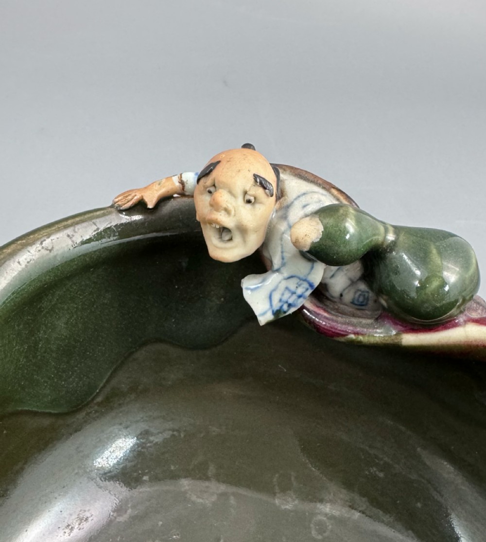 A Japanese Sumida Gawa pottery bowl featuring a dark green running glaze over a red base with two sculpted figures leaning over the rim and an applied white makers mark. View V