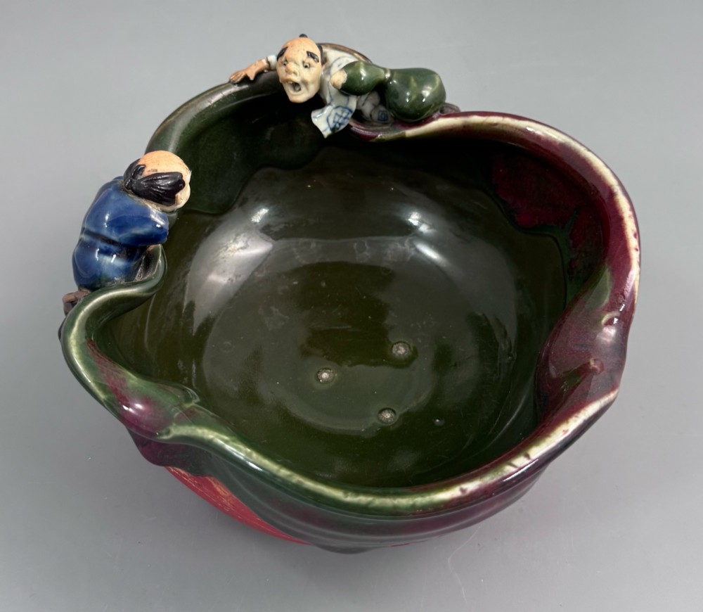 A Japanese Sumida Gawa pottery bowl featuring a dark green running glaze over a red base with two sculpted figures leaning over the rim and an applied white makers mark. View VI