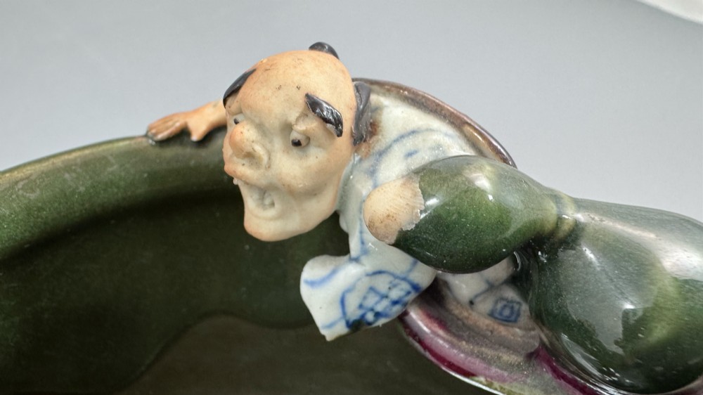 A Japanese Sumida Gawa pottery bowl featuring a dark green running glaze over a red base with two sculpted figures leaning over the rim and an applied white makers mark. View VII