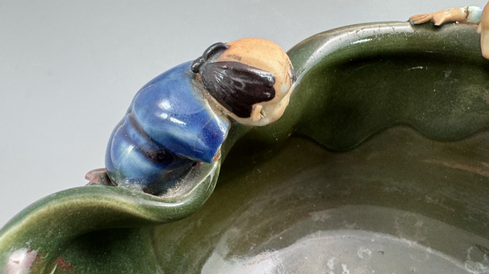 A Japanese Sumida Gawa pottery bowl featuring a dark green running glaze over a red base with two sculpted figures leaning over the rim and an applied white makers mark. View VIII
