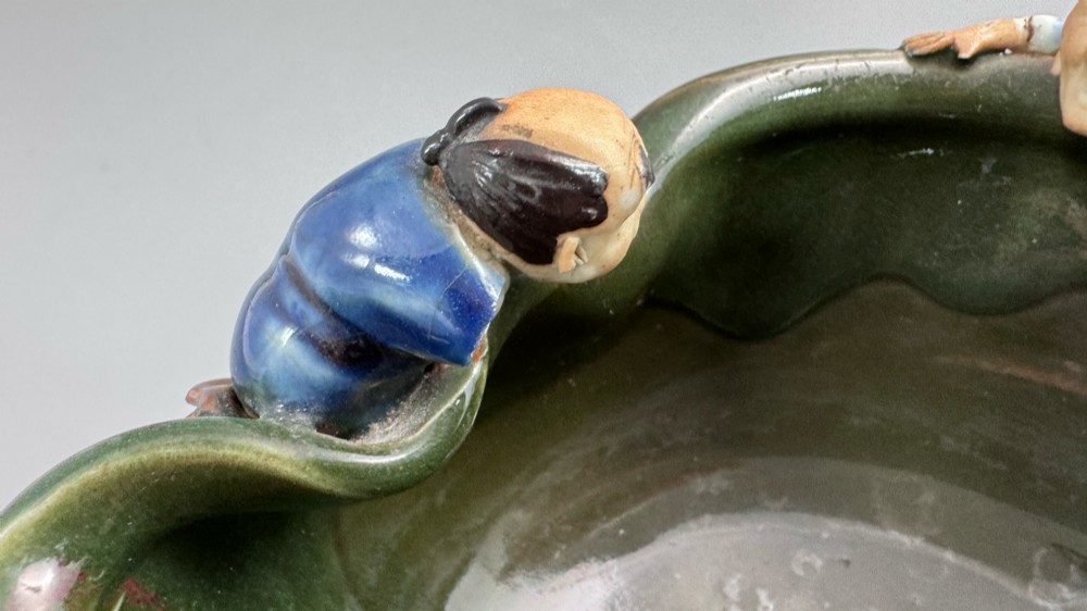 A Japanese Sumida Gawa pottery bowl featuring a dark green running glaze over a red base with two sculpted figures leaning over the rim and an applied white makers mark. View IX
