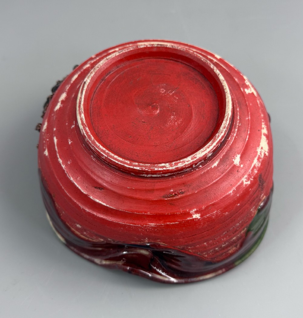 A Japanese Sumida Gawa pottery bowl featuring a dark green running glaze over a red base with two sculpted figures leaning over the rim and an applied white makers mark. View X