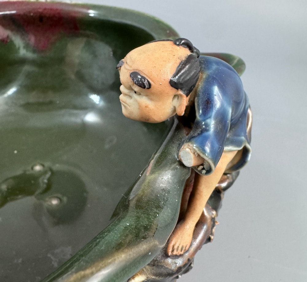 A Japanese Sumida Gawa pottery bowl featuring a dark green running glaze over a red base with two sculpted figures leaning over the rim and an applied white makers mark. View XIV