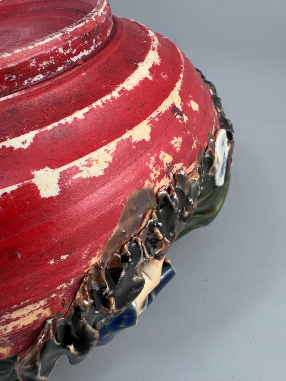 A Japanese Sumida Gawa pottery bowl featuring a dark green running glaze over a red base with two sculpted figures leaning over the rim and an applied white makers mark. View XV