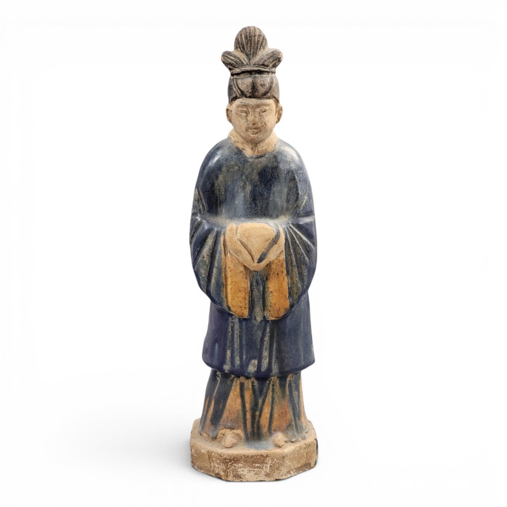 A Chinese Tang dynasty Sancai glazed earthenware figure of a court attendant standing on a hexagonal base with hands clasped under draped sleeves.