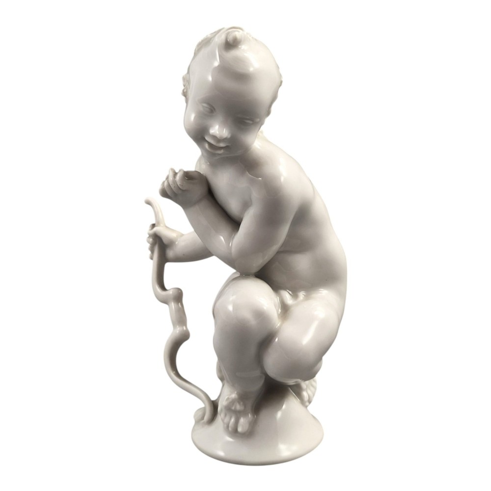 A glazed white porcelain figurine depicting a crouching winged cherub holding a curved bow on a circular base. View II