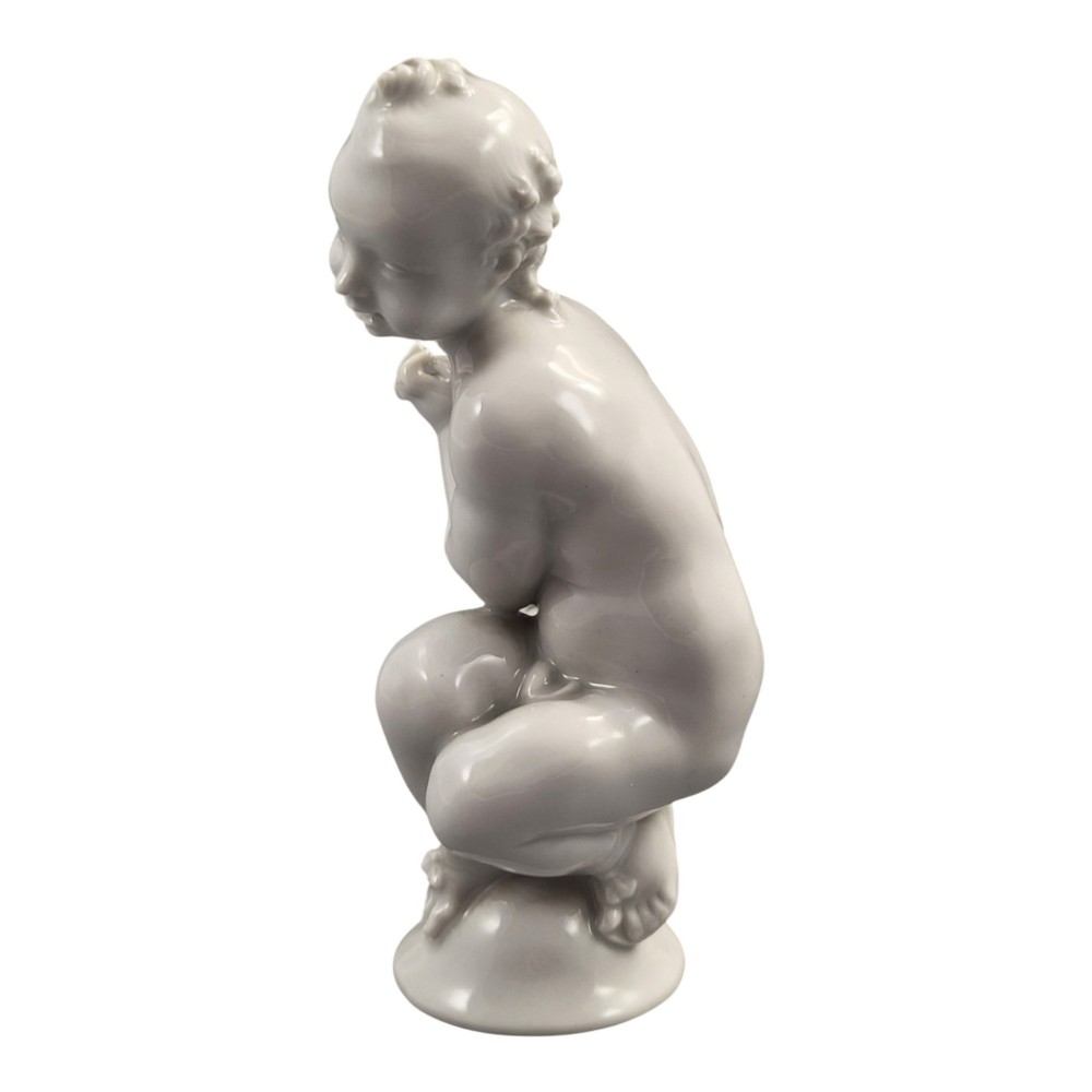 A glazed white porcelain figurine depicting a crouching winged cherub holding a curved bow on a circular base. View III