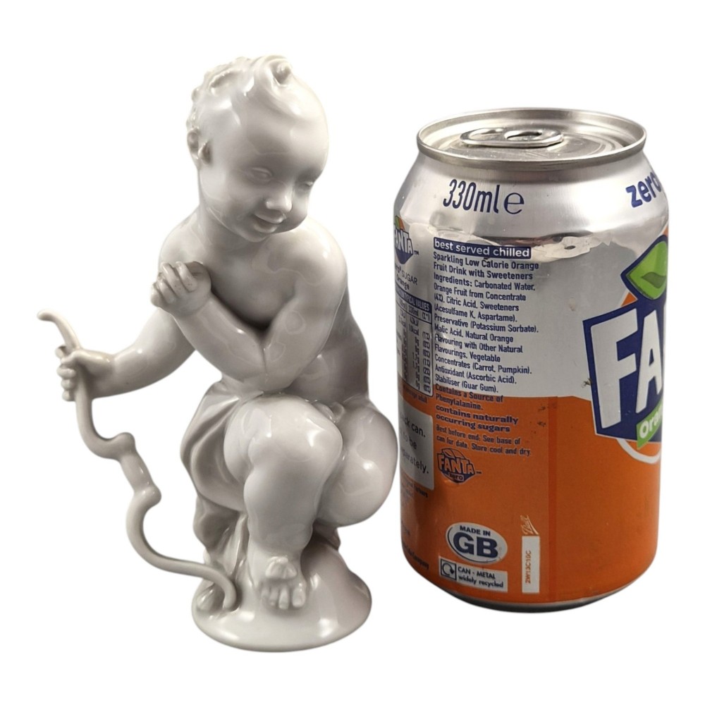 A glazed white porcelain figurine depicting a crouching winged cherub holding a curved bow on a circular base. View X