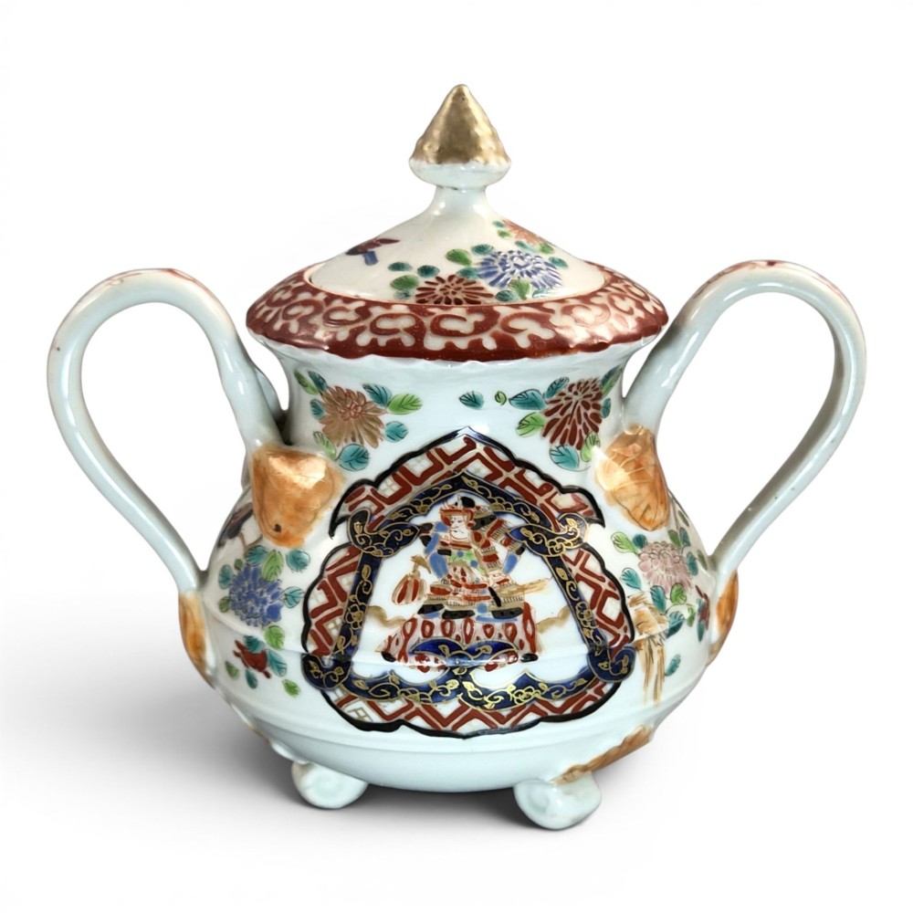 A covered Japanese Imari porcelain sugar bowl featuring double side handles, a pointed gilded finial, and hand-painted samurai and floral motifs in red, blue, green, and gold enamels.