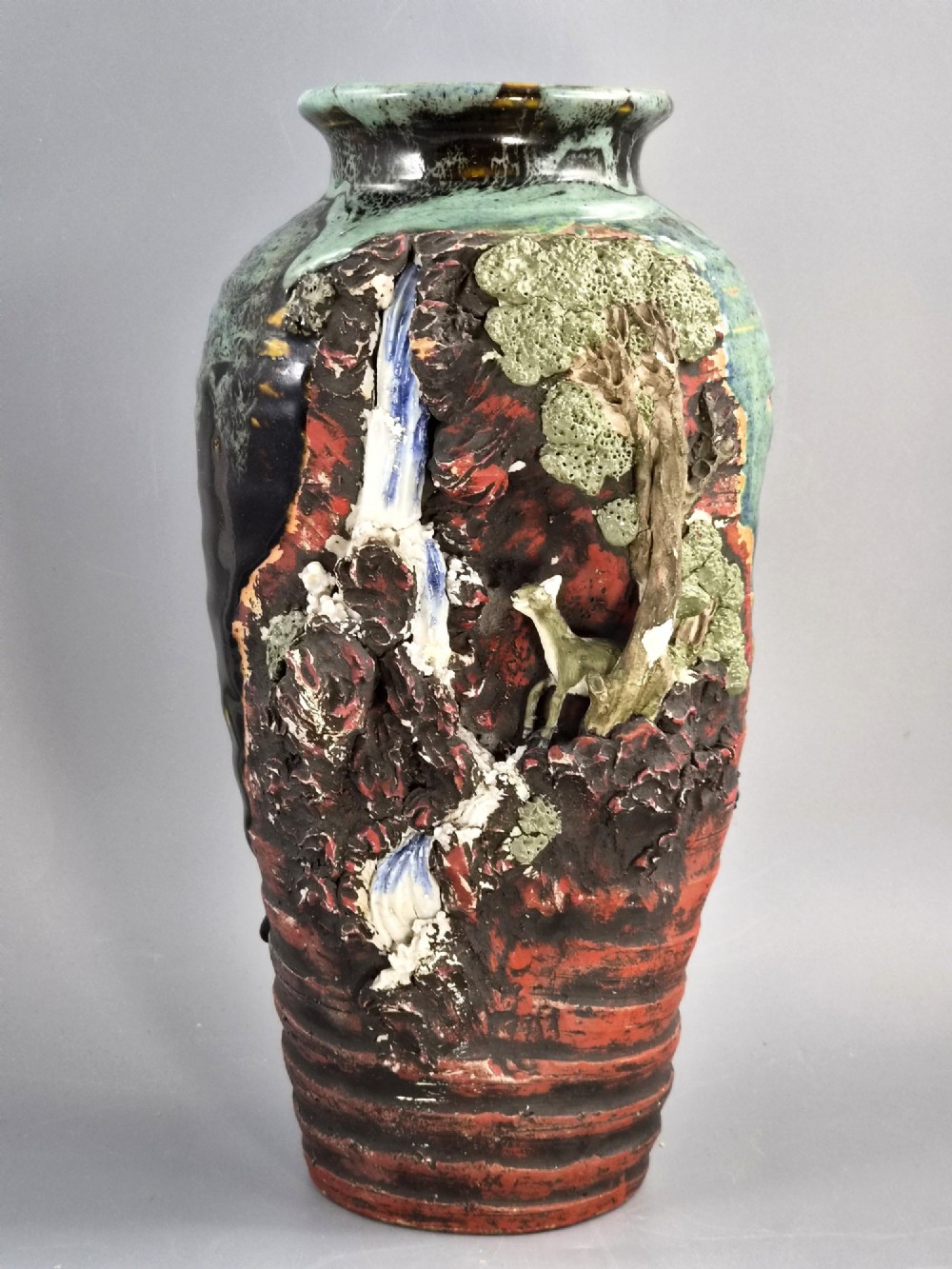 A Japanese Sumida Gawa stoneware vase featuring high-relief sculptures of a stag, waterfall, and tree against a ribbed dark red base with a green drip glaze top rim.