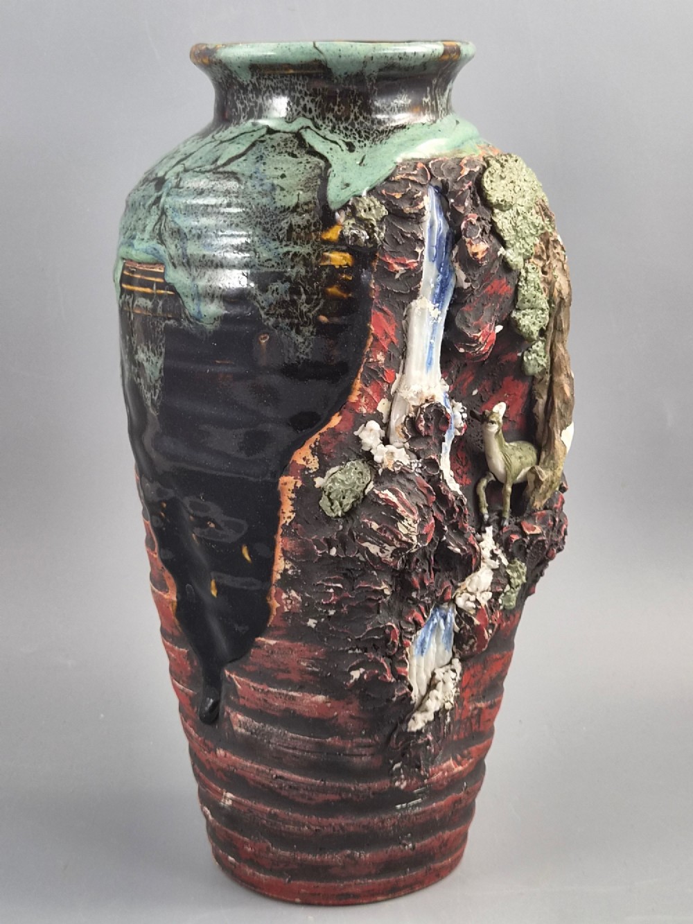 A Japanese Sumida Gawa stoneware vase featuring high-relief sculptures of a stag, waterfall, and tree against a ribbed dark red base with a green drip glaze top rim. View VI