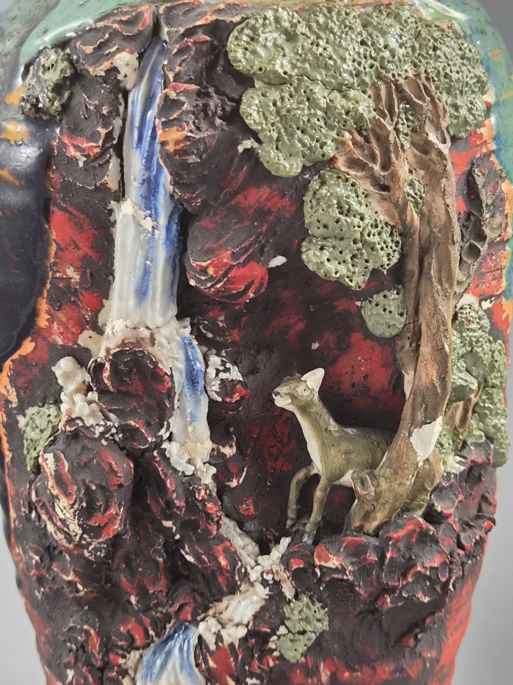 A Japanese Sumida Gawa stoneware vase featuring high-relief sculptures of a stag, waterfall, and tree against a ribbed dark red base with a green drip glaze top rim. View VII