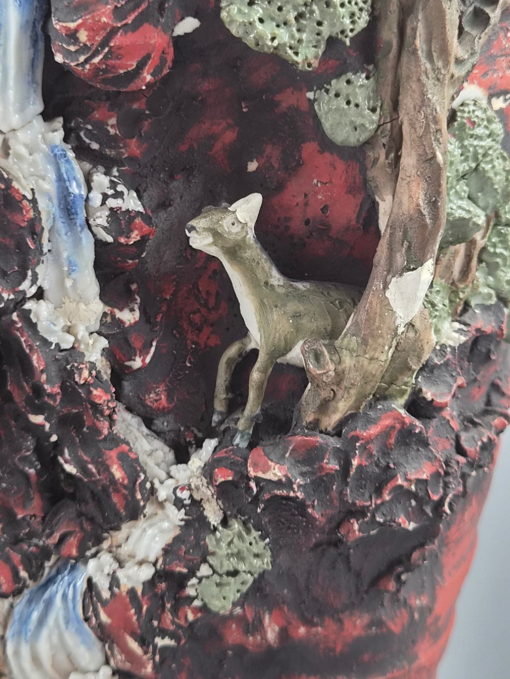 A Japanese Sumida Gawa stoneware vase featuring high-relief sculptures of a stag, waterfall, and tree against a ribbed dark red base with a green drip glaze top rim. View VIII
