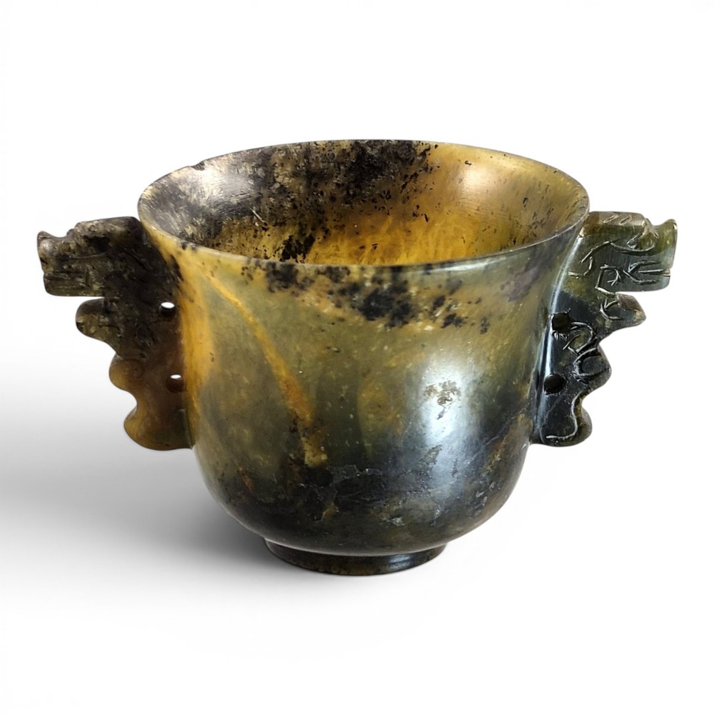 A hand-carved mottled olive-green and dark ochre nephrite jade cup featuring carved handles on either side.