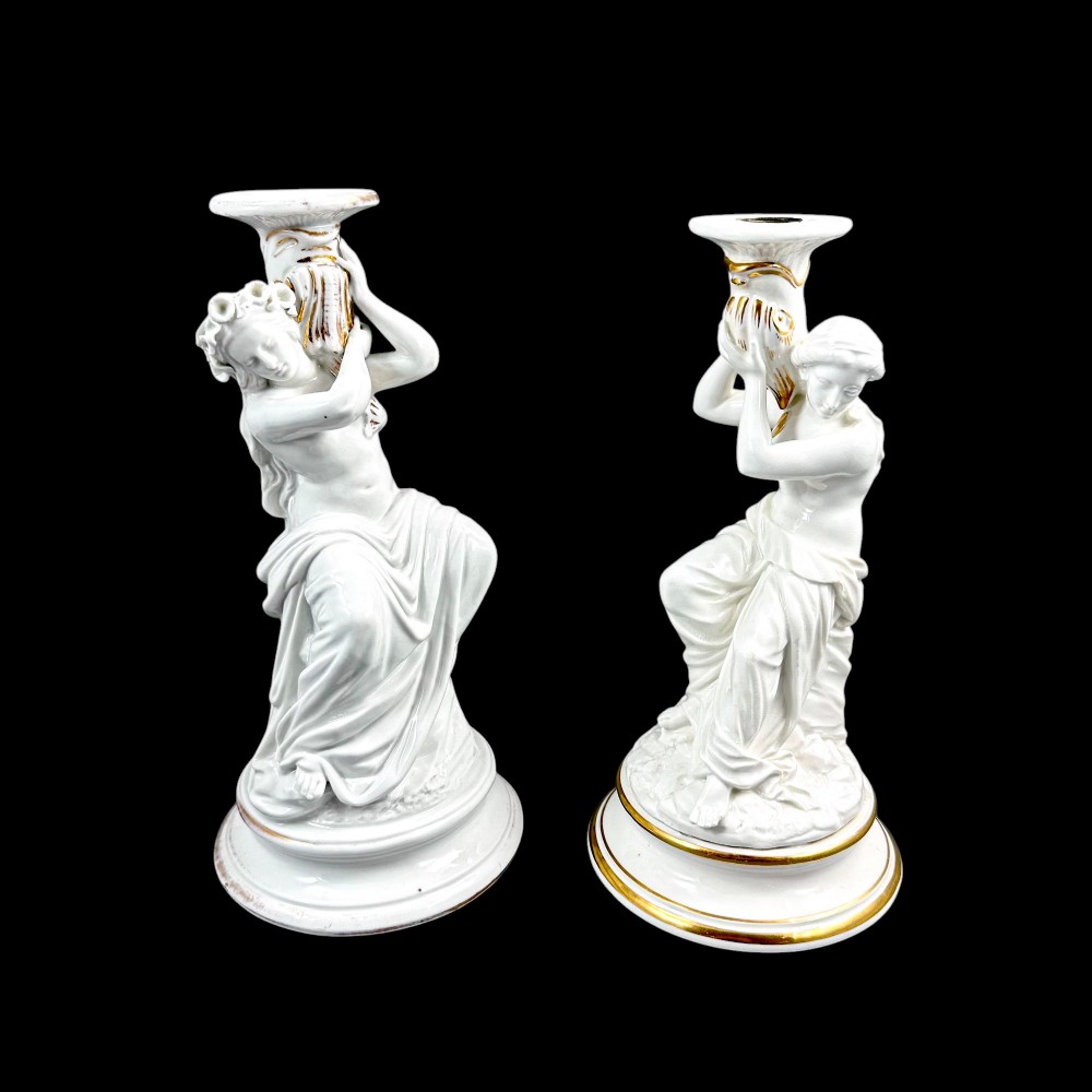A pair of white glazed porcelain candlesticks featuring sculpted classical female figures holding urn-shaped sconces with gilt trim on circular bases.