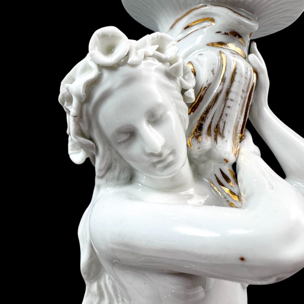 A pair of white glazed porcelain candlesticks featuring sculpted classical female figures holding urn-shaped sconces with gilt trim on circular bases. View V