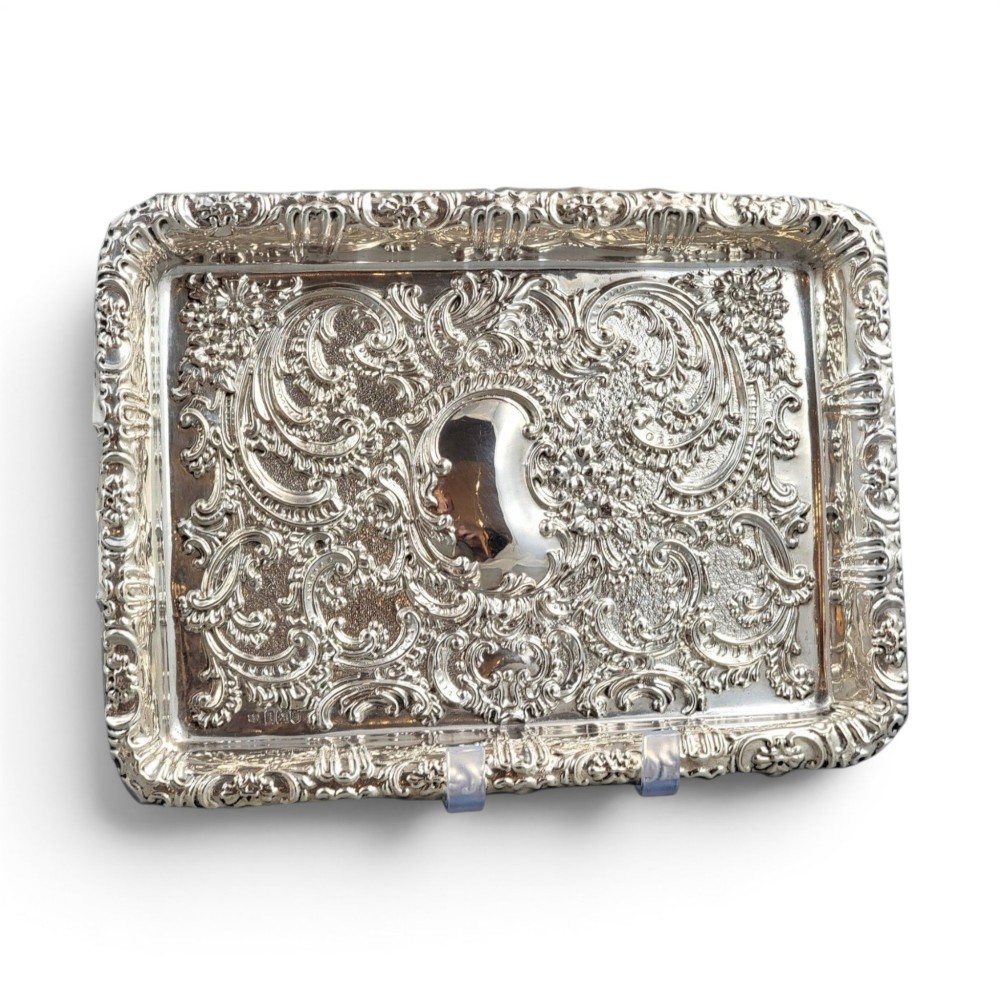 A rectangular Edwardian sterling silver card tray with intricate floral repousse scrollwork detailing, a plain central cartouche, and a raised scalloped rim.