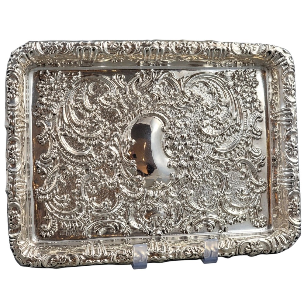 A rectangular Edwardian sterling silver card tray with intricate floral repousse scrollwork detailing, a plain central cartouche, and a raised scalloped rim. View II
