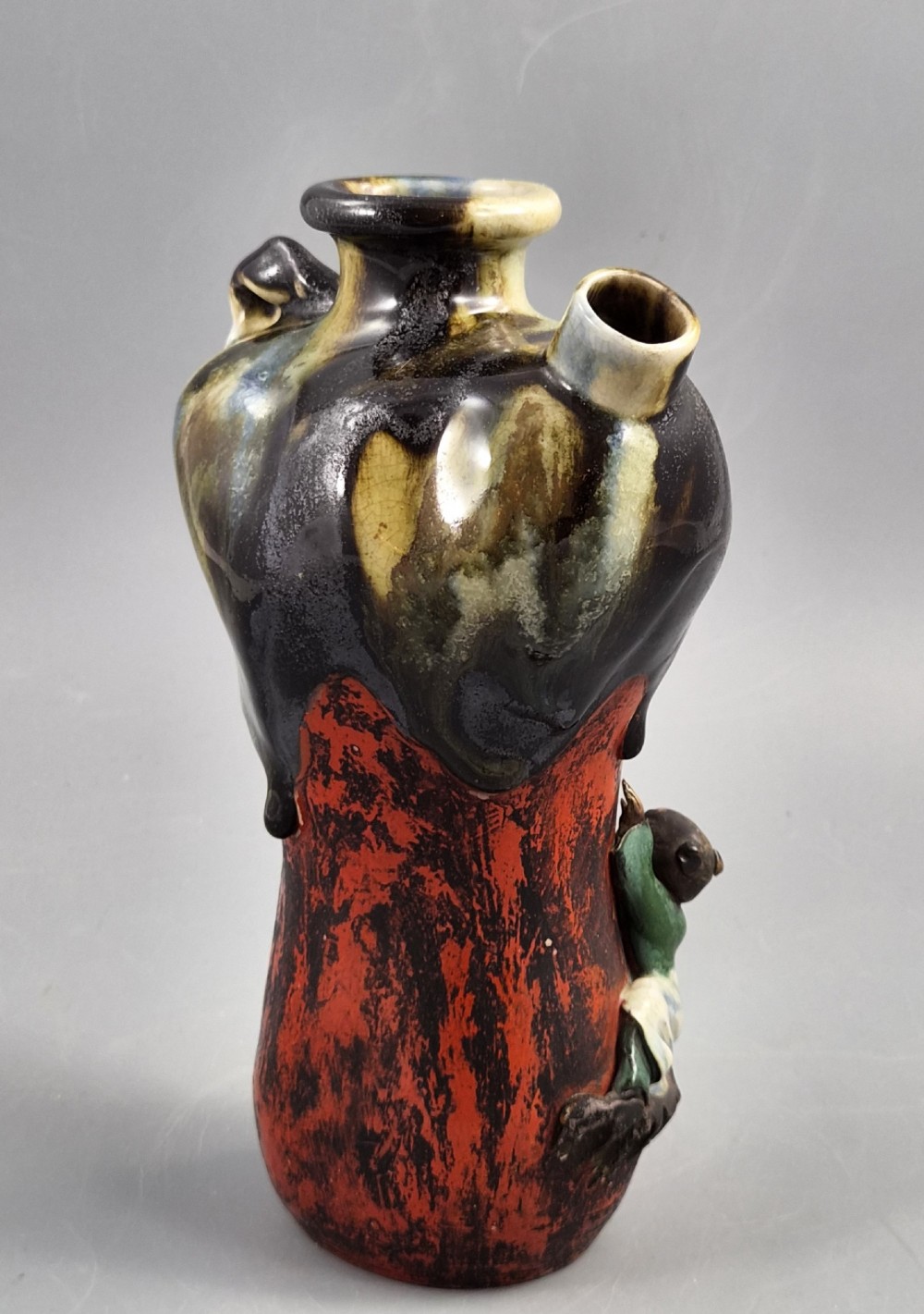 A Japanese Sumida Gawa pottery vase featuring an asymmetrical spouted form with a dark flambe drip glaze over a textured red lower body adorned with applied high-relief figures of boys. View VI