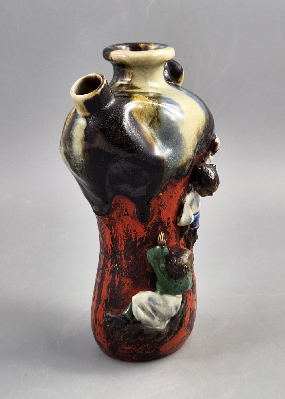 A Japanese Sumida Gawa pottery vase featuring an asymmetrical spouted form with a dark flambe drip glaze over a textured red lower body adorned with applied high-relief figures of boys. View VII