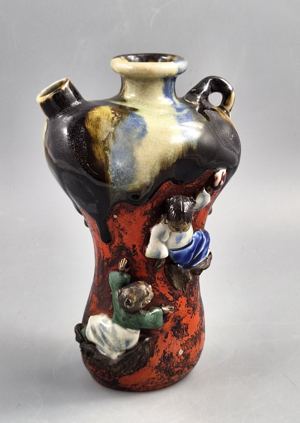 A Japanese Sumida Gawa pottery vase featuring an asymmetrical spouted form with a dark flambe drip glaze over a textured red lower body adorned with applied high-relief figures of boys. View VIII