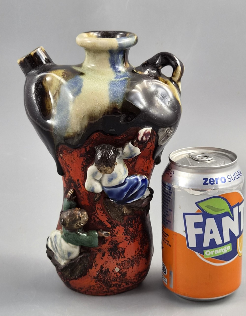 A Japanese Sumida Gawa pottery vase featuring an asymmetrical spouted form with a dark flambe drip glaze over a textured red lower body adorned with applied high-relief figures of boys. View XI