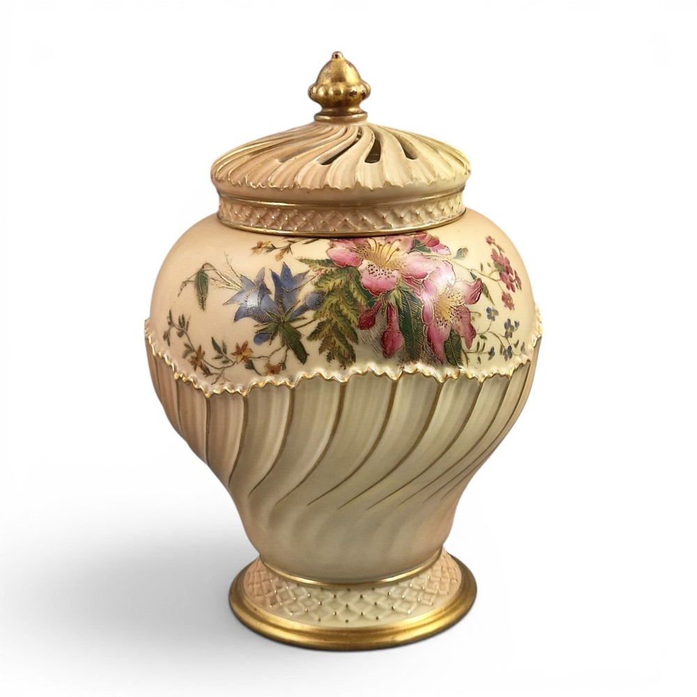 A Royal Worcester blush ivory porcelain pot-pourri jar with a pierced lid, fluted swirled lower body, gold trim, and hand-painted floral motifs.