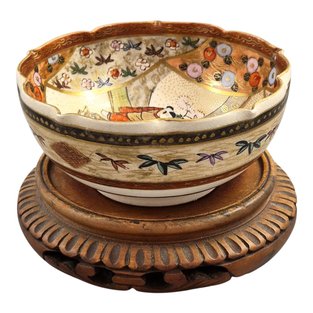 A small lobed Japanese Satsuma ceramic bowl with intricate polychrome floral painting and gold gilding, resting on a carved dark wooden base. View III