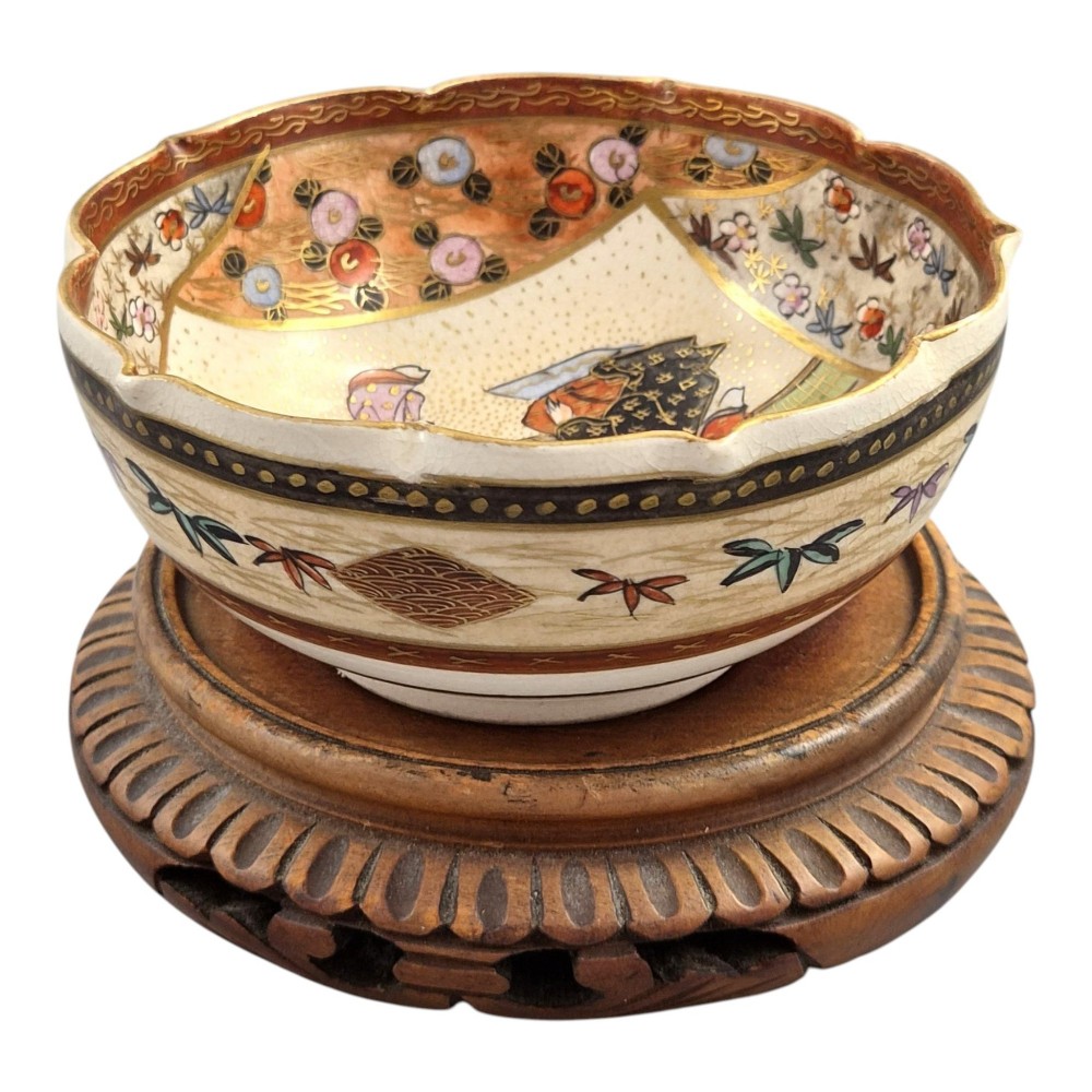 A small lobed Japanese Satsuma ceramic bowl with intricate polychrome floral painting and gold gilding, resting on a carved dark wooden base. View V