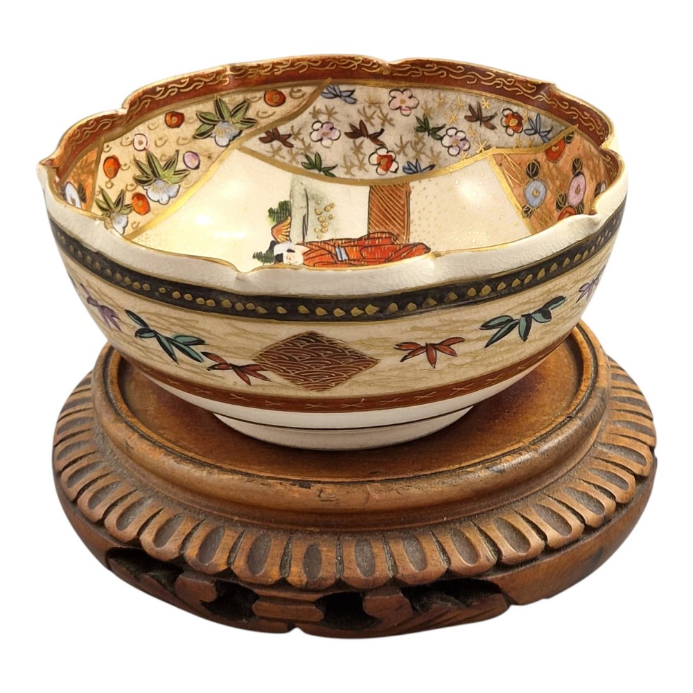 A small lobed Japanese Satsuma ceramic bowl with intricate polychrome floral painting and gold gilding, resting on a carved dark wooden base. View VI
