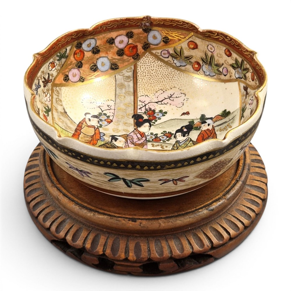 A small lobed Japanese Satsuma ceramic bowl with intricate polychrome floral painting and gold gilding, resting on a carved dark wooden base. View X