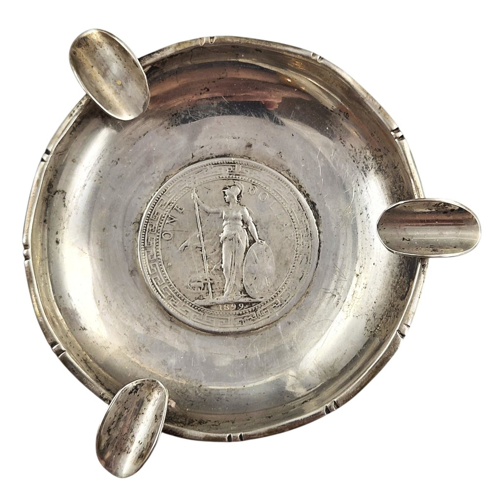 A circular sterling silver dish featuring three cigarette rests on the outer rim and an inset 1899 British Trade Dollar coin in the centre.