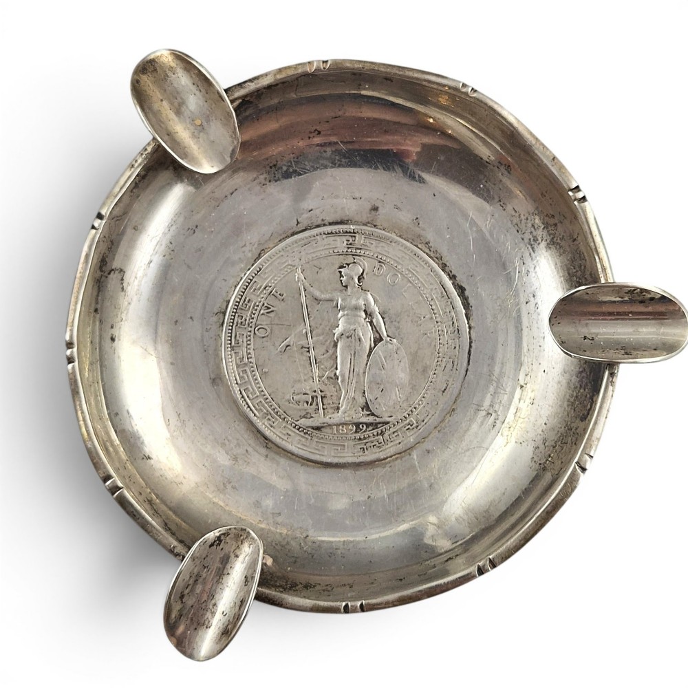 A circular sterling silver dish featuring three cigarette rests on the outer rim and an inset 1899 British Trade Dollar coin in the centre. View II