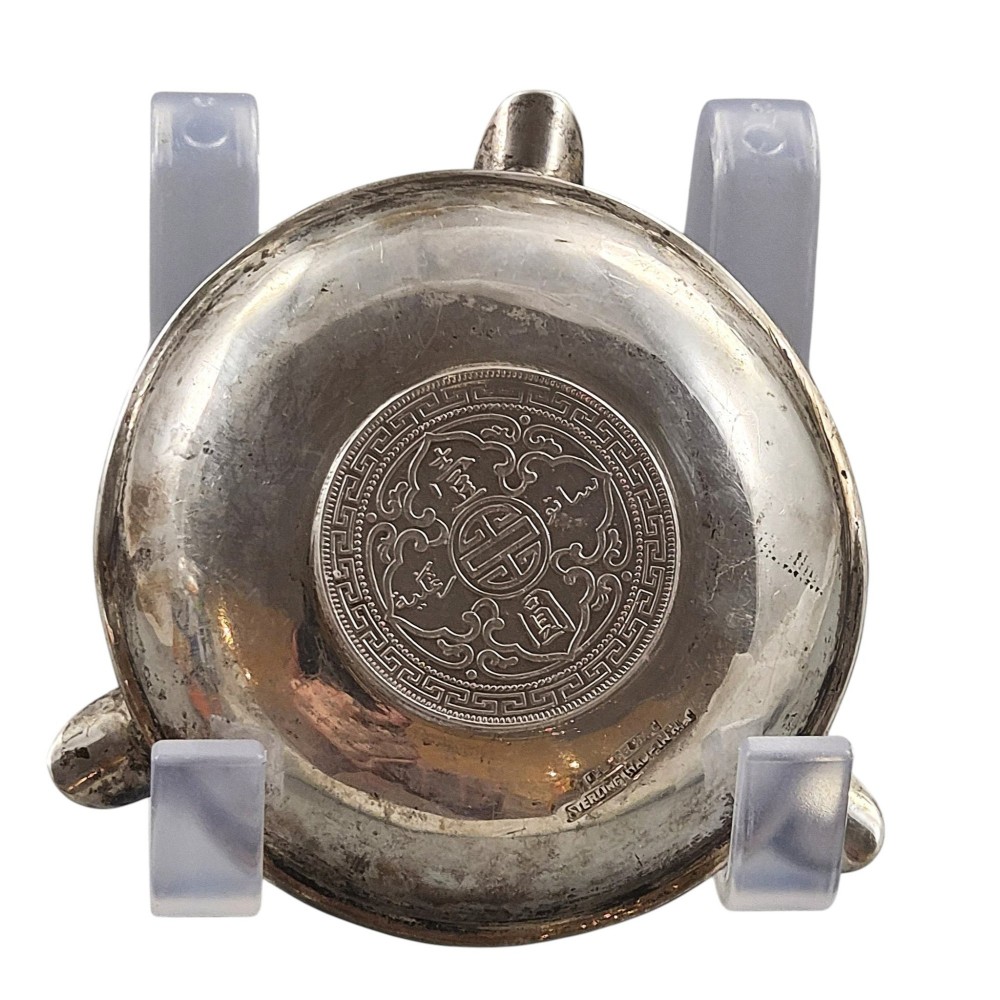 A circular sterling silver dish featuring three cigarette rests on the outer rim and an inset 1899 British Trade Dollar coin in the centre. View IV